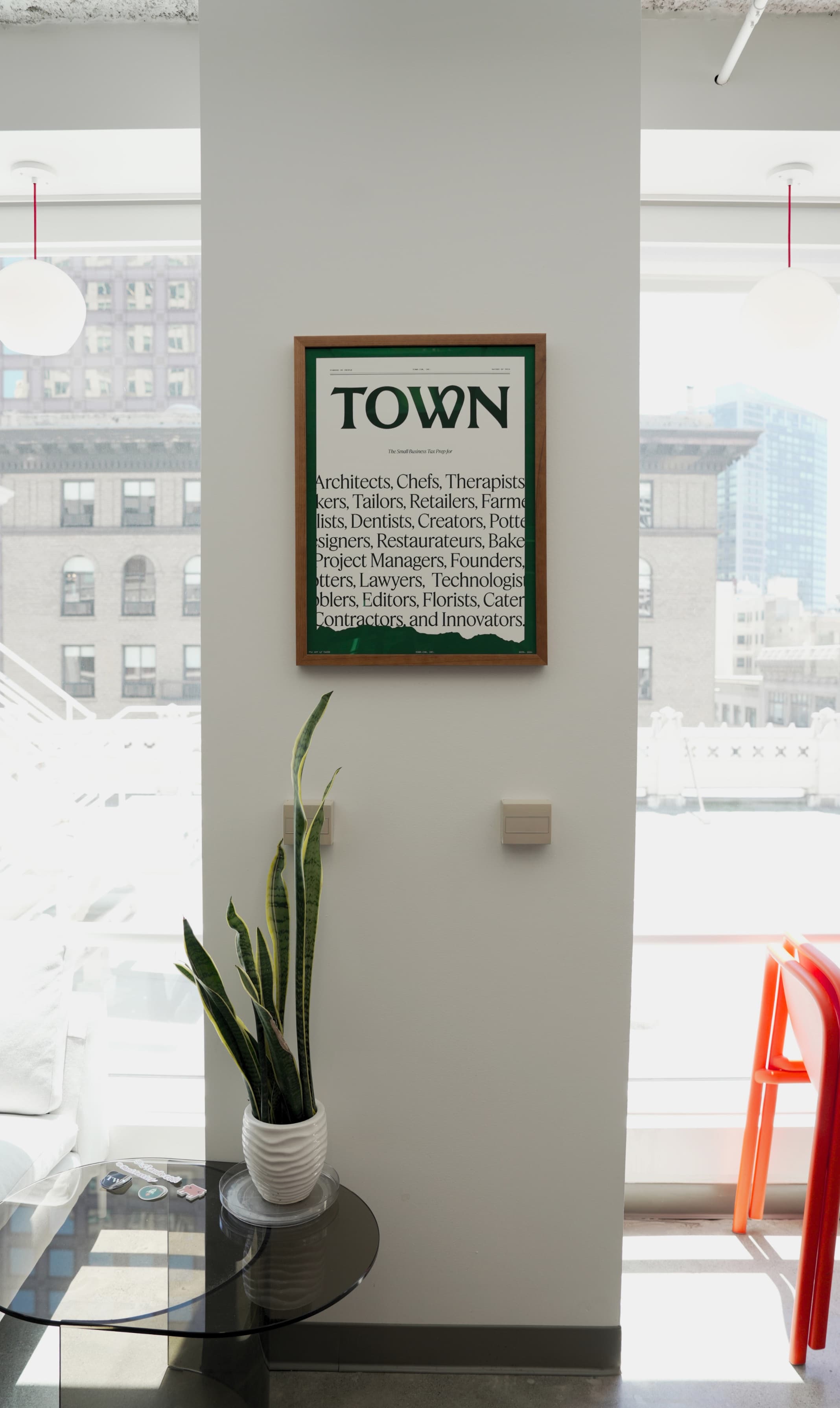 A framed Town print on a wall in Town's office — "TOWN" above a list of the people Town is for — beside a plant and city windows.